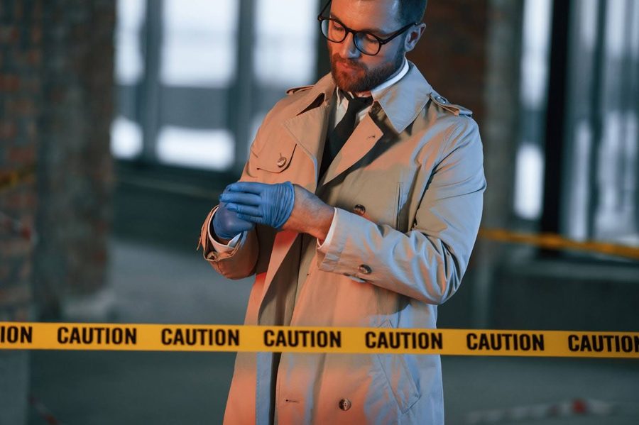 An investigator in a trench coat pulls on a latex glove behind yellow caution tape at a scene.