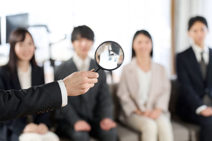A magnifying glass hovers over a seated group, symbolizing candidate screening.