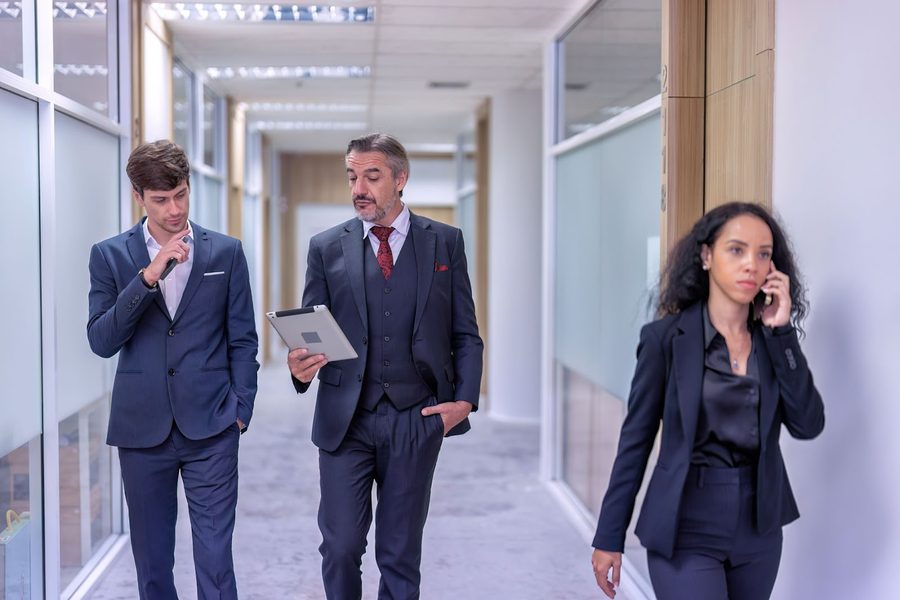 Two men in suits walk down an office corridor reviewing a tablet while a woman on a phone call passes them.