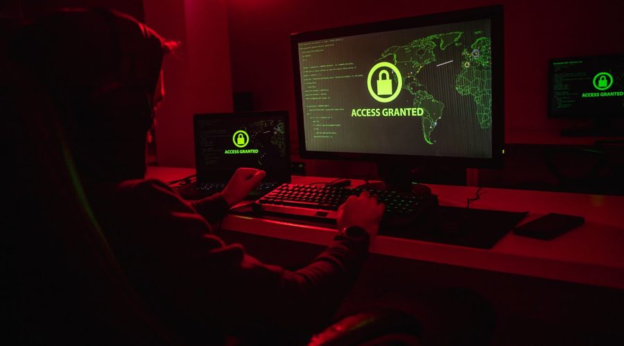 An anonymous hacker is stealing data on a computer in red lighting.