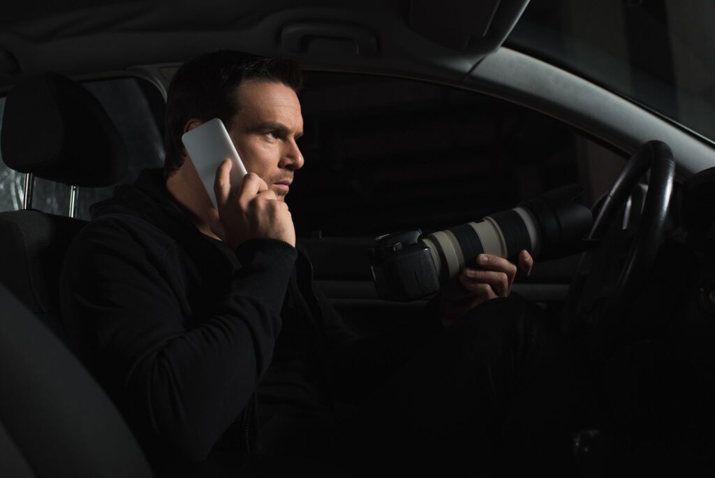 A private investigator talking on his smartphone while taking photographs with a camera at night from inside his car.