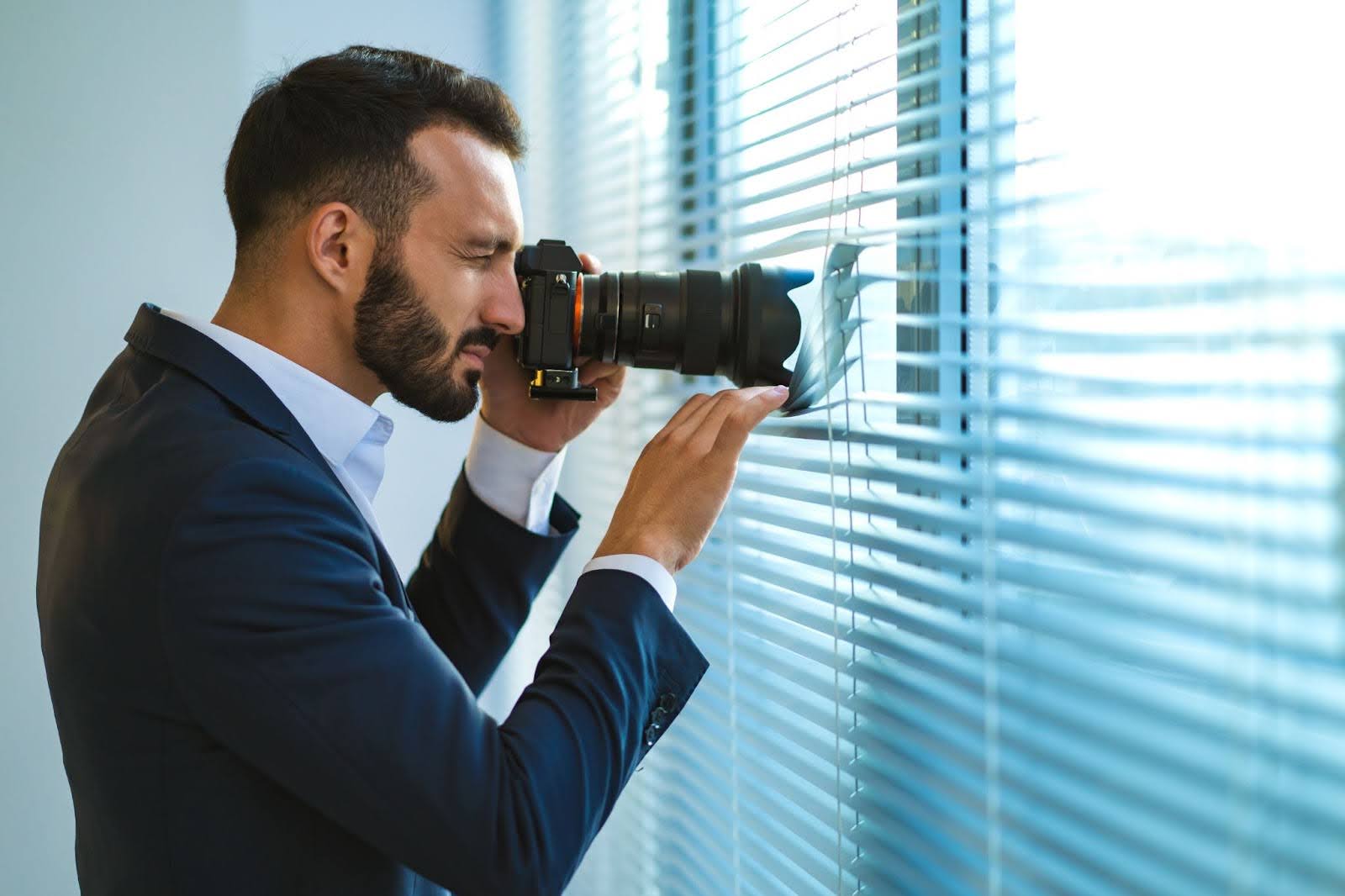 A man dressed in a suit sticks a camera through blinds to take discreet photos.