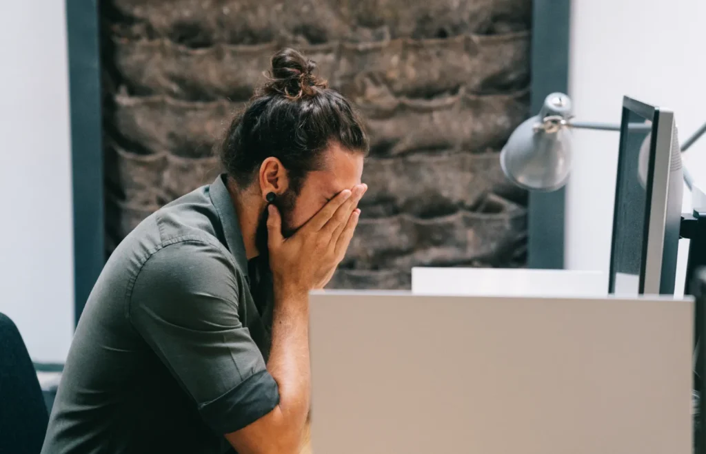 An emotional man puts his head in his hands in front of his computer screen.