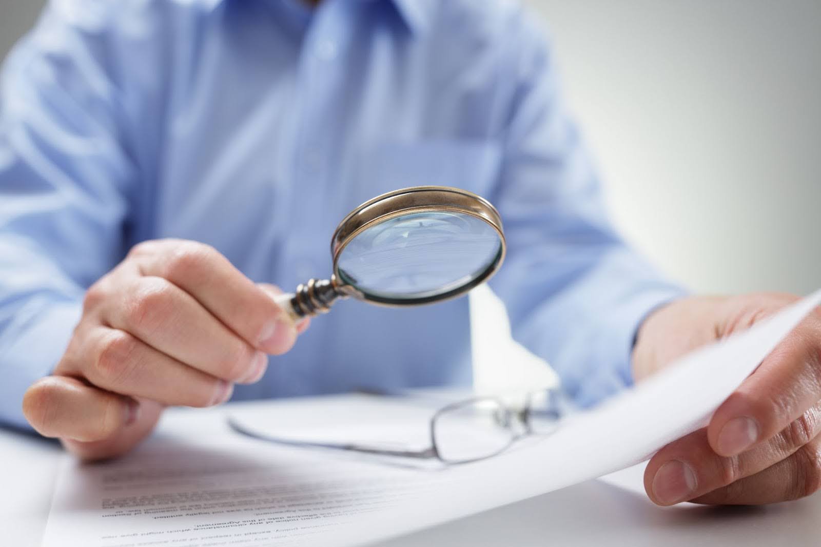 Insurance investigator examining a document with a magnifying glass beside his eyeglasses.